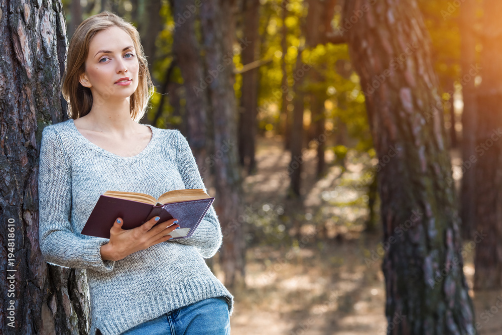 Obraz premium Beautiful blond girl resting in spring or autumn forest read book and standing. Confident caucasian young woman in casual clothes relaxing near big tree in park and holding book. Attractive female