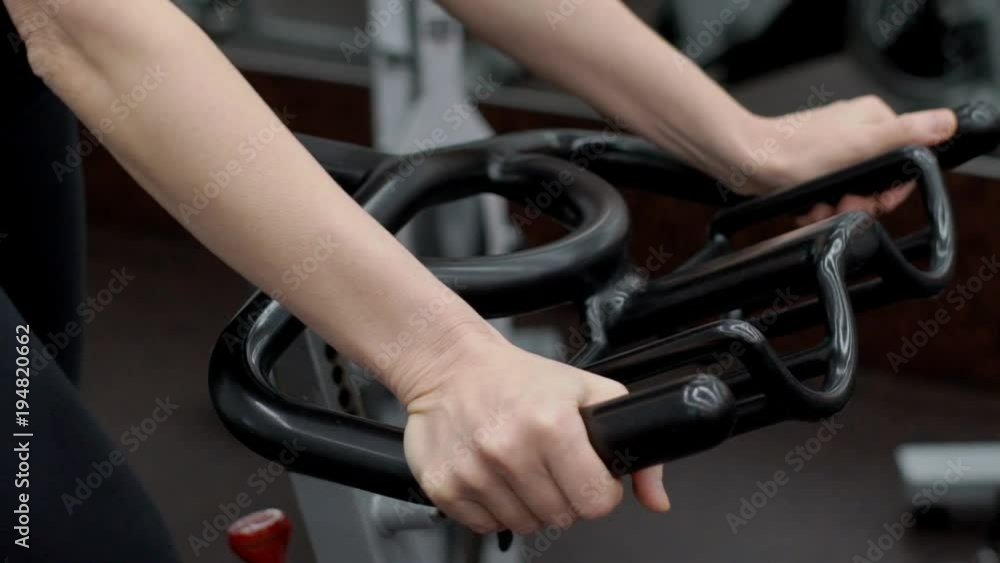 Young woman is holding the black handlebar of stationary bicycle in modern gym, close up. Lady in sportswear is having a training and doing exercises to meke her ggluteus and harmstrings stronger and Young woman is holding the black handlebar of stationary bicycle in modern gym, close up. Lady in sportswear is having a training and doing exercises to meke her ggluteus and harmstrings stronger and