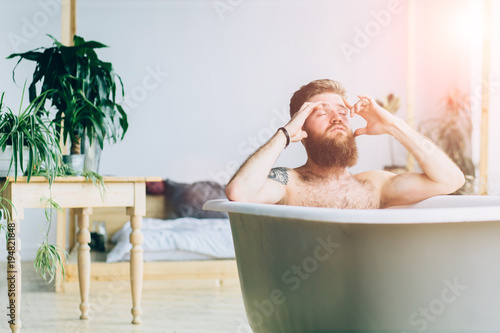 Man with beard meditation, relaxing and having a bath in a white buthtub in loft interior studio. photo