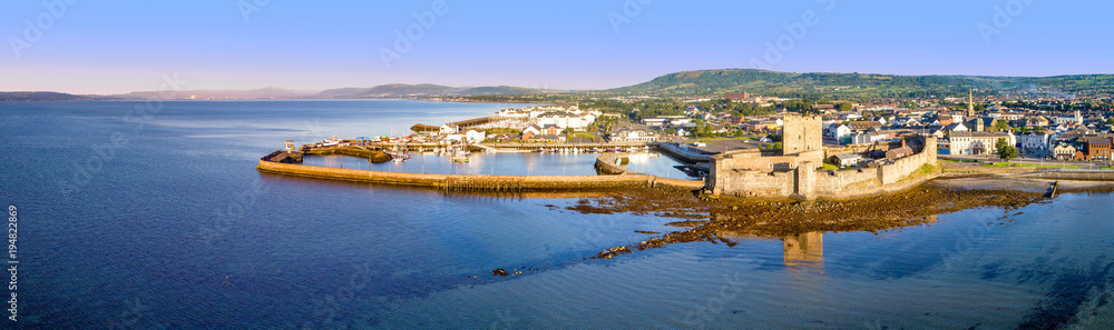 Medieval Norman Castle in Carrickfergus near Belfast in sunrise light. Aerial wide panorama with marina, yachts, parking, town, Belfast Lough and far view of Belfast city in the background.