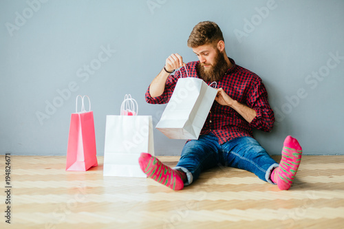 Hipster beard man relaxing after shopping. Male sitting at floor at home with shopping bags over gray wall background.