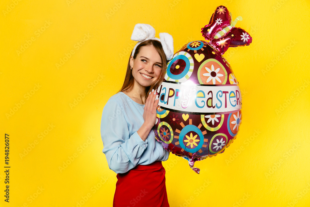 Easter. Holiday concept. Gorgeous woman wearing bunny ears, holding egg shape balloon, celebrating Easter Day. Dressed in colorful clothes, isolated on yellow. Easter. Holiday concept. Gorgeous woman wearing bunny ears, holding egg shape balloon, celebrating Easter Day. Dressed in colorful clothes, isolated on yellow.