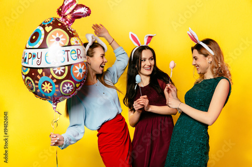 Easter. Happy Easter Day. Attractive young women holding big air balloon, colorful eggs, enjoying the holiday. Isolated on yellow background. Dressed in vivid clothes.