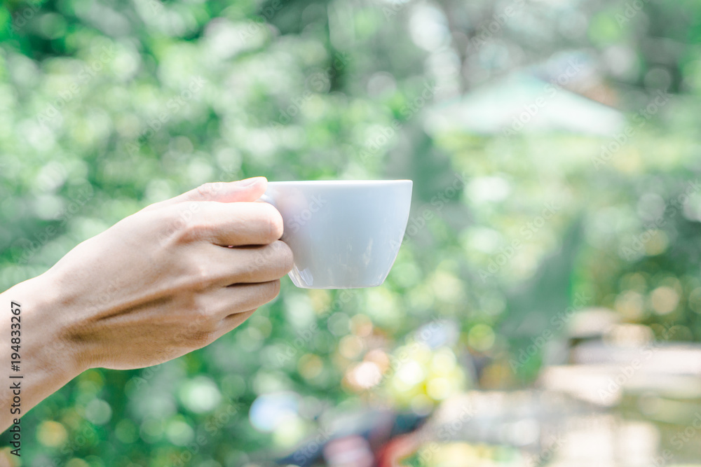 Hand holding white cup of coffee over blur on many leaves background, mock up, template, green leaves concept Hand holding white cup of coffee over blur on many leaves background, mock up, template, green leaves concept