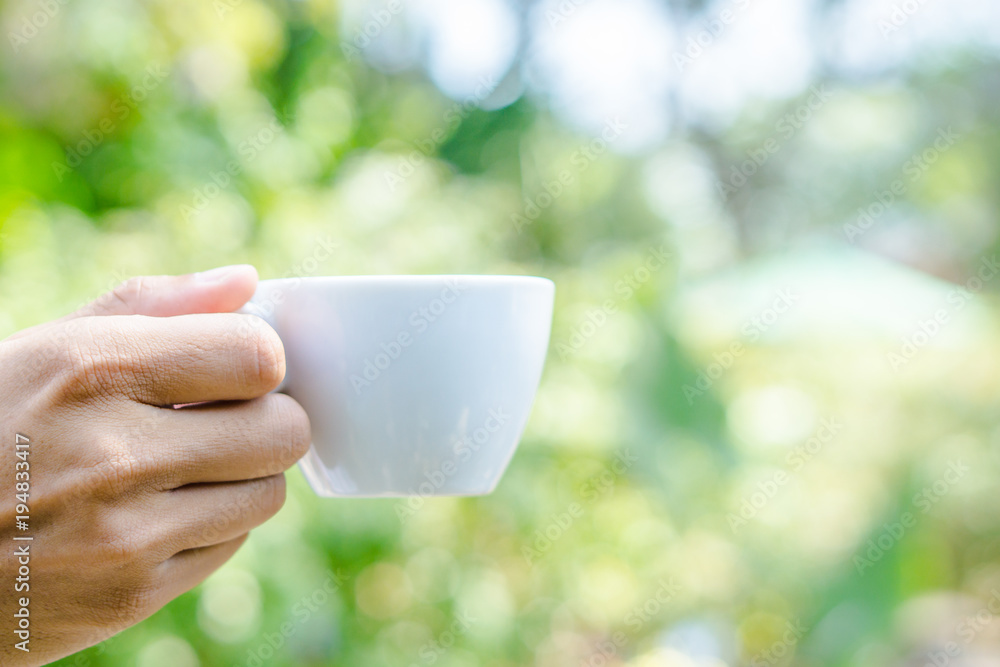 Hand holding white cup of coffee over blur on many leaves background, mock up, template, green leaves concept Hand holding white cup of coffee over blur on many leaves background, mock up, template, green leaves concept
