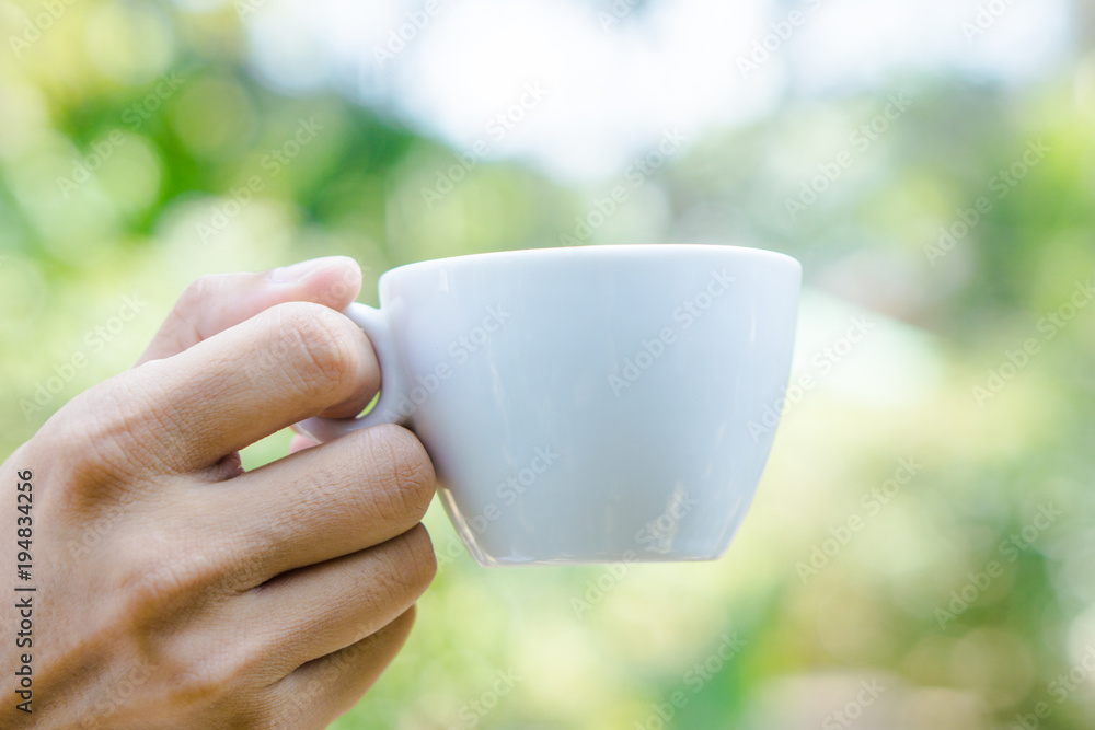 Hand holding white cup of coffee over blur on many leaves background, mock up, template, green leaves concept Hand holding white cup of coffee over blur on many leaves background, mock up, template, green leaves concept