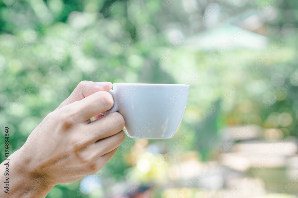 Hand holding white cup of coffee over blur on many leaves background, mock up, template, green leaves concept Hand holding white cup of coffee over blur on many leaves background, mock up, template, green leaves concept