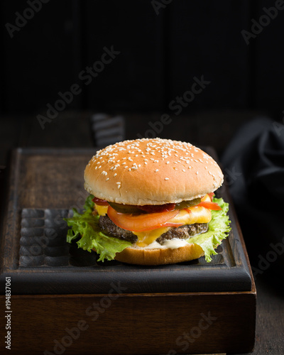 Delicious fresh tasty burger with beef, tomato, cheese and lettuce served on a wooden cutting board on dark background. Street fast food