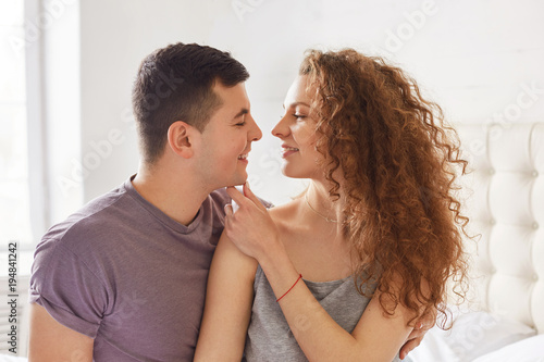 Portrait of affectionate lovely couple: curly woman and brunet male look at each other`s eyes with great love, feel closeness and togetherness, enjoy calm domestic atmopshere. Romantic relations