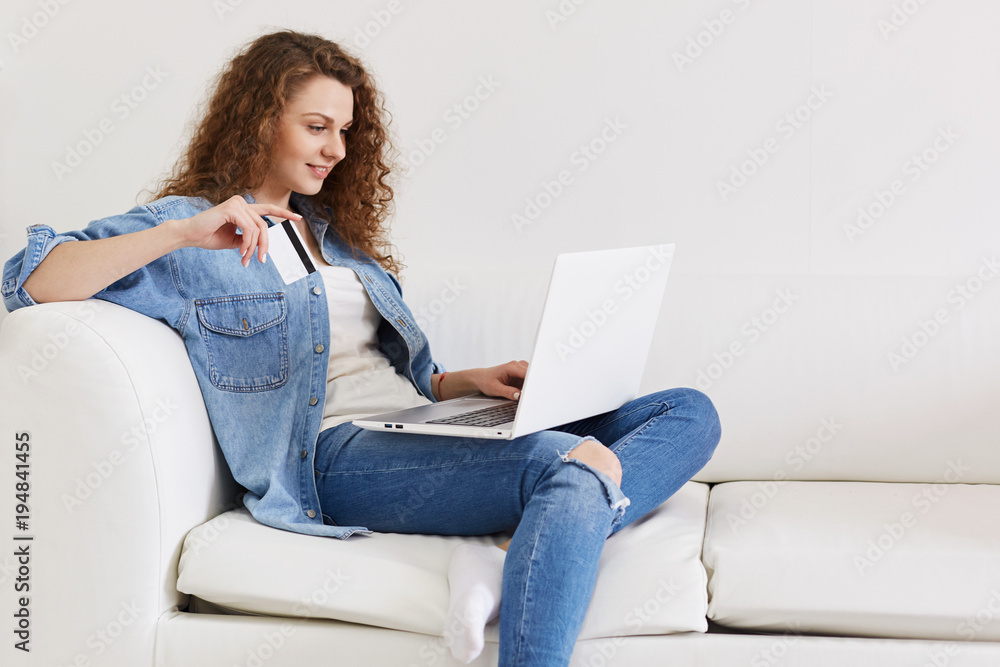 Pleased young curly female model in stylish denim clothing holds plastic card and portable laptop computer, surf internet webstores and makes shopping online, rejoices new successful purchases Pleased young curly female model in stylish denim clothing holds plastic card and portable laptop computer, surf internet webstores and makes shopping online, rejoices new successful purchases