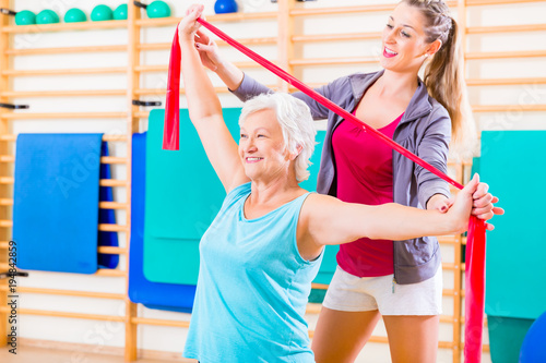 Senior woman with stretch band in fitness gym being coached by personal trainer