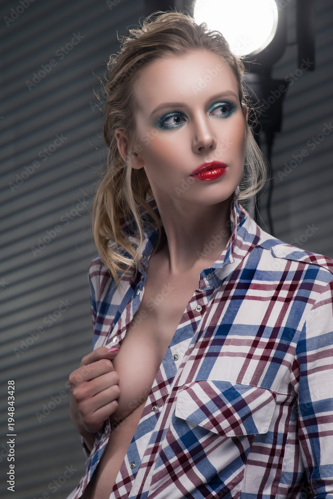 Beautiful slender blonde big breast girl without a bra wearing unbuttoned  checkered shirt and jeans on a striped gray background near the studio  lamp. Stock Photo | Adobe Stock