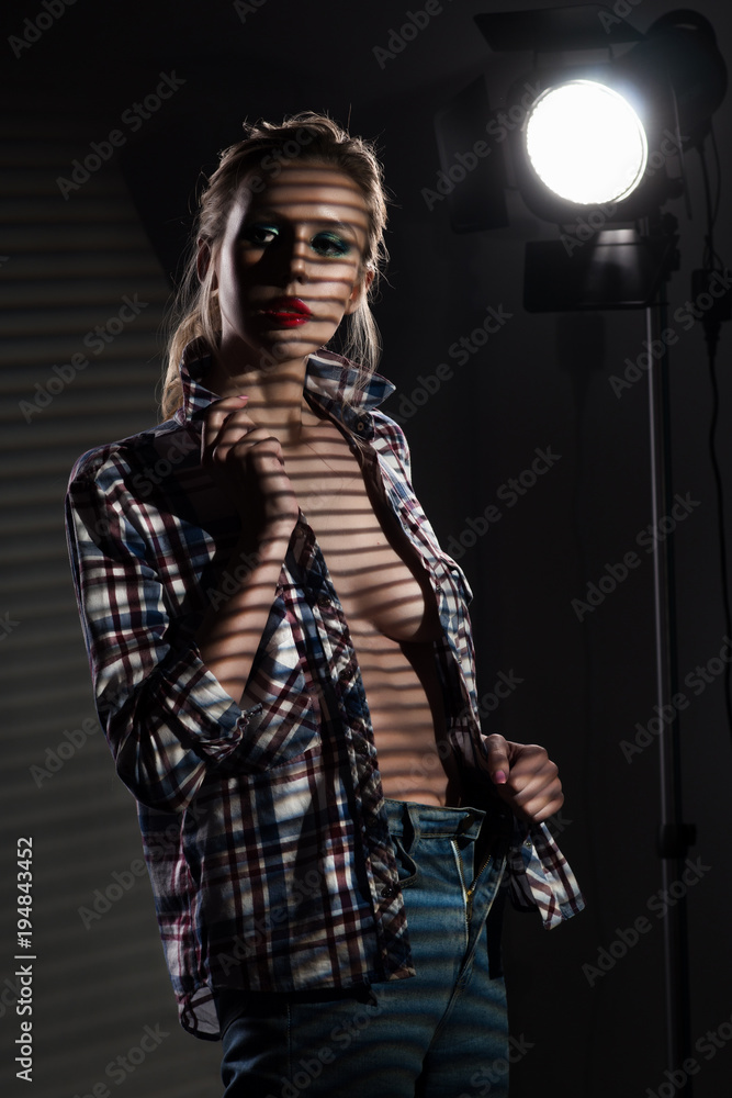 Naklejka premium A beautiful slender blonde big breasts girl without a bra wearing an unbuttoned checkered shirt and jeans covered with shadows from the blinds near the studio lamp