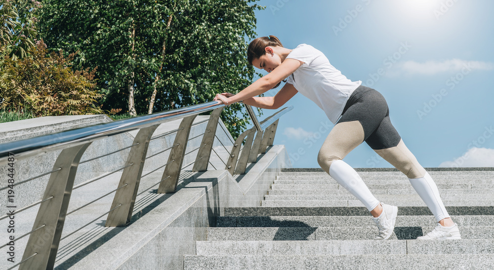 Sunny summer day. Young woman in sportswear doing stretching exercises outdoor. Girl doing warm-up on steps before training. Exercise in street, sports exercises, workout. On background blue sky.