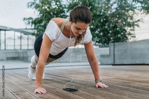 Summer day. Young woman doing sports exercises outdoors. Girl makes plank while looking at smartphone screen and listening to music. Workout, sports, sports training.