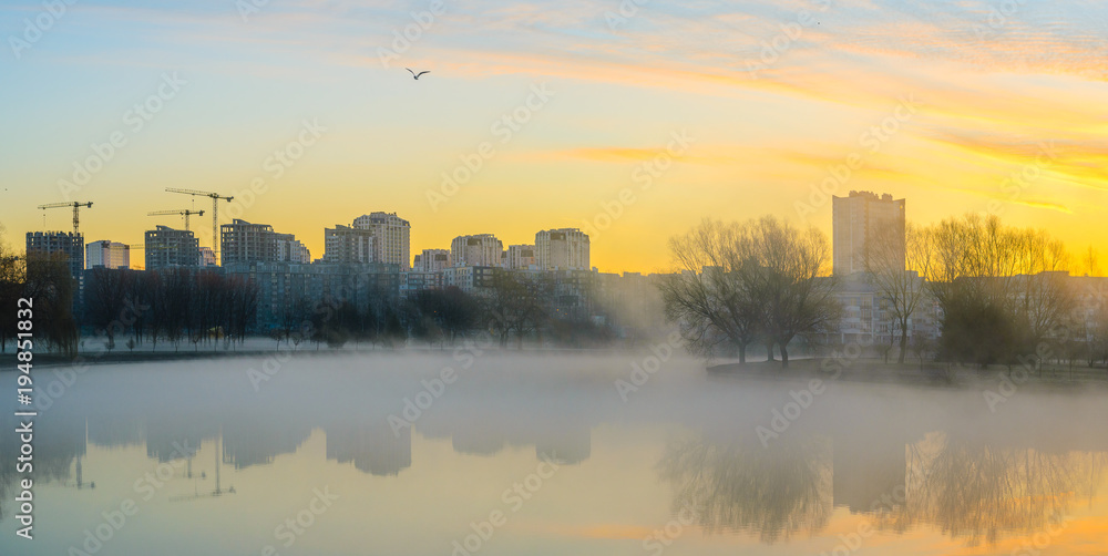 Heavy fog on water in morning. City skyline with multistory buildings on river bank