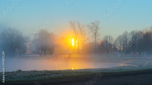 A lone silhouette of a cyclist riding in a strong fog along the river bank in a city park early in the morning. Rays of the sun in the reflection of the windows of buildings cut through a thick fog