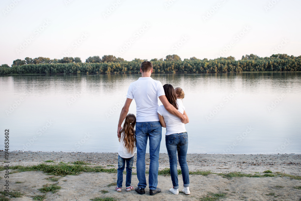 young happy couple with children standing embracing on the river bank. Happy young father hugs his wife and two daughters young happy couple with children standing embracing on the river bank. Happy young father hugs his wife and two daughters