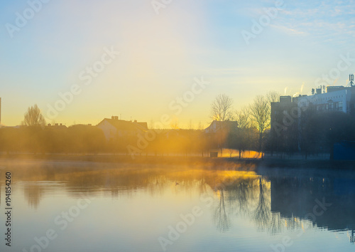 Weather conditions: a strong morning fog on a pond near residential buildings. Reflection of the facades of residential buildings at the surface of the water at dawn. Abstract weather background
