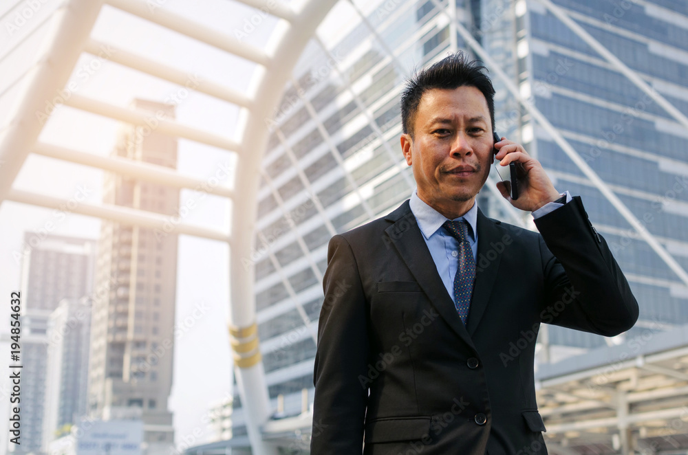 medium aged asian business man wearing modern black suit making phone call with mobile smart phone in building city background, internet network connection, technology communication, financial concept medium aged asian business man wearing modern black suit making phone call with mobile smart phone in building city background, internet network connection, technology communication, financial concept