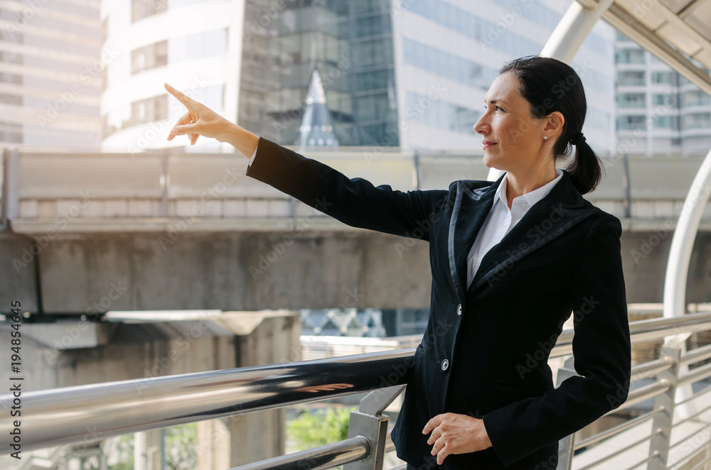 medium aged smart caucasian business woman wearing modern black suit pointing up and looking up to future standing in building city background, confident, team leader, successful and manager concept medium aged smart caucasian business woman wearing modern black suit pointing up and looking up to future standing in building city background, confident, team leader, successful and manager concept