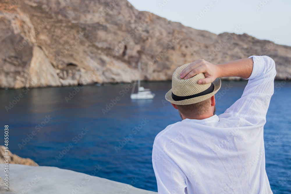 A man in a hat and white shirt is sitting with his back on the seashore, and staring at the yacht