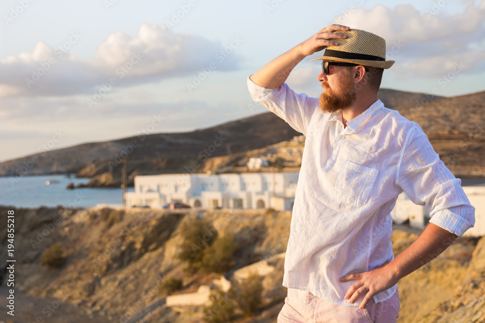 Red-bearded male traveler in a white shirt stands in the rays of the morning sun on a steep bank near the sea