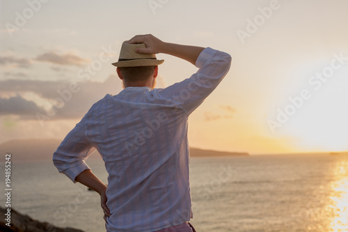 Silhouette of a man in white clothes and a hat stands with his back in the golden rays of the sun against the background of a dawn over the sea