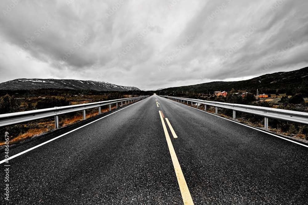 Fototapeta premium Remote road leading through the wilderness of Norway with asphalt , forest and mountains on a dark cloudy sky