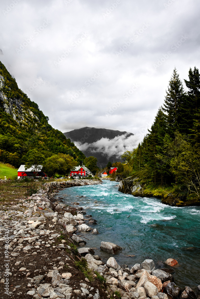 Red scandinavian wooden houses and cabins at a wild and fast turquoise river and shoreline surrounded by mountains and thick forest and trees in Norway