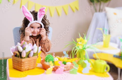 baby girl in rabbit ears sitting at table on background of Easter decor