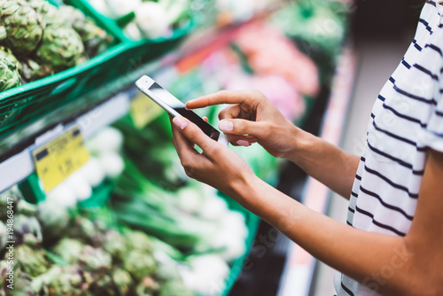 woman shopping purchase healthy food in supermarket blur background, view girl blogger buy products using smartphone in store. Hipster at grocery using smartphone. Person comparing price