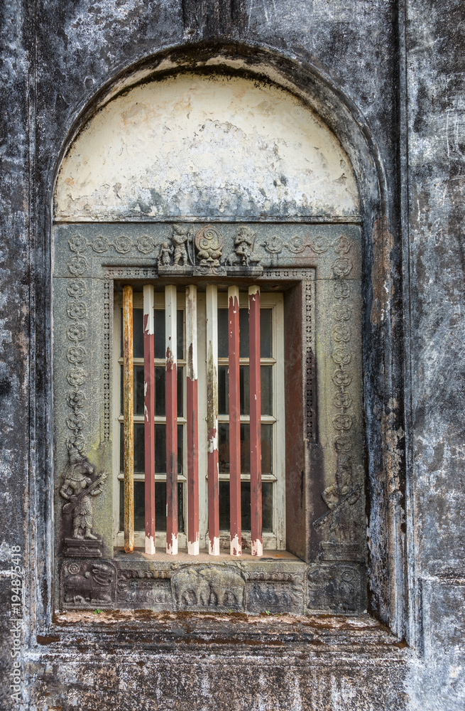Madikeri, India - October 31, 2013: Closeup of window and rusty decorated metal frame of Royal mausoleum, set in white wall devastated by black mold. Hindu figurines.