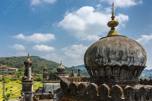 Madikeri, India - October 31, 2013: Domes, golden flags and turrets on top of two Royal mausolea at Raja tomb domain under blue sky. Closeup of one dome in front. Green hills in back. Shot on roof. photo