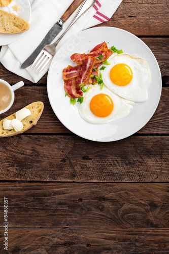 Fried eggs, bacon and italian ciabatta bread on white plate. Cup of coffee. Breakfast. Top view. Wooden background