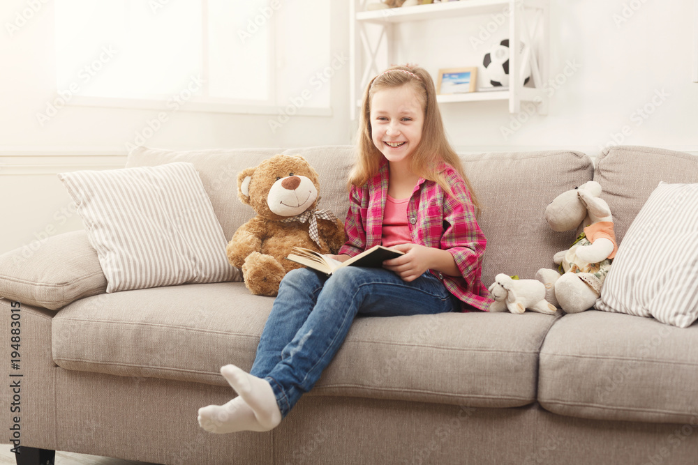 Happy little female child hugging her teddy bear and reading book on sofa at home