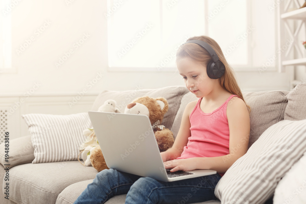 Cute little girl doing homework on laptop, sitting on couch in headphones Cute little girl doing homework on laptop, sitting on couch in headphones