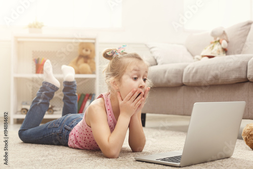 Scared casual little girl watching movie on laptop while lying on the floor at home © Prostock-studio