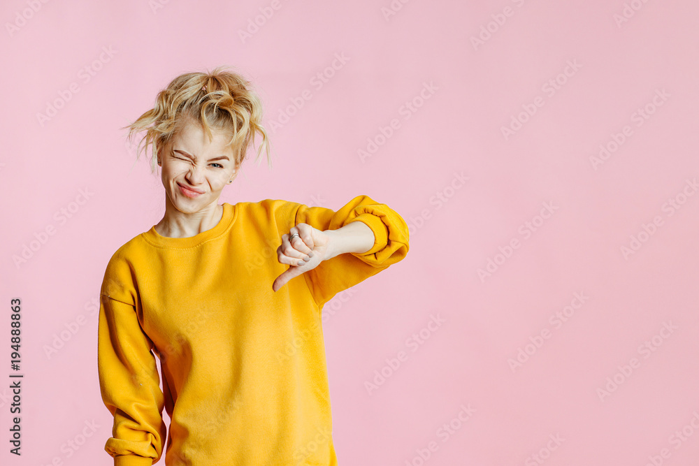 close-up portrait of cheerful young caucasian female with curly blonde hair, in yellow sweater poses on a pink background. woman making thumb down, dissatisfied and angry. close-up portrait of cheerful young caucasian female with curly blonde hair, in yellow sweater poses on a pink background. woman making thumb down, dissatisfied and angry.
