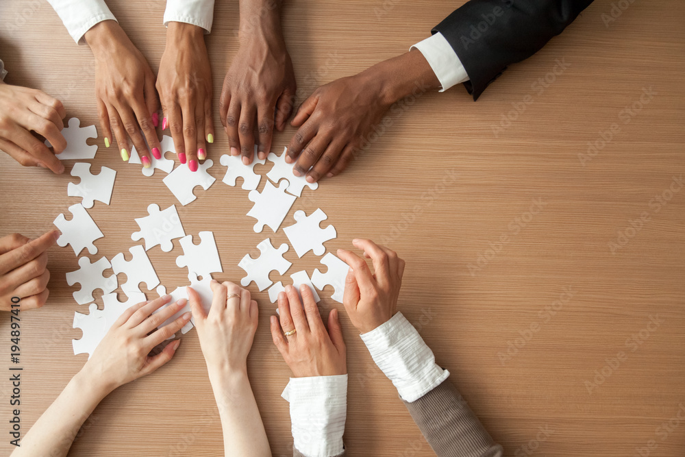 Hands of multi-ethnic team assembling jigsaw puzzle, multiracial group of  black and white people joining pieces at desk, successful teamwork concept,  help and support in business, close up top view Stock Photo