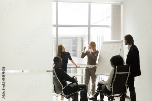 Diverse business team listening to female speaker discussing presentation on flip chart at meeting in boardroom, group brainstorm and marketing sales coaching corporate training concept, copy space