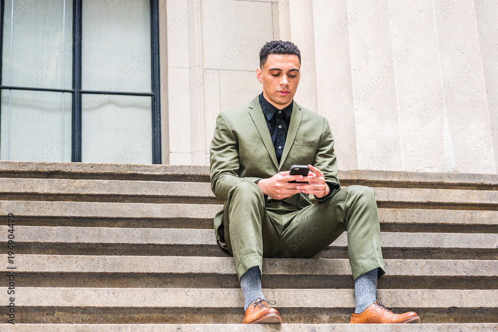 Young American Businessman traveling, working in New York, wearing green suit, brown leather shoes, siting on stairs outside office on street, taking work break, looking down, texting on cell phone. . Young American Businessman traveling, working in New York, wearing green suit, brown leather shoes, siting on stairs outside office on street, taking work break, looking down, texting on cell phone. .