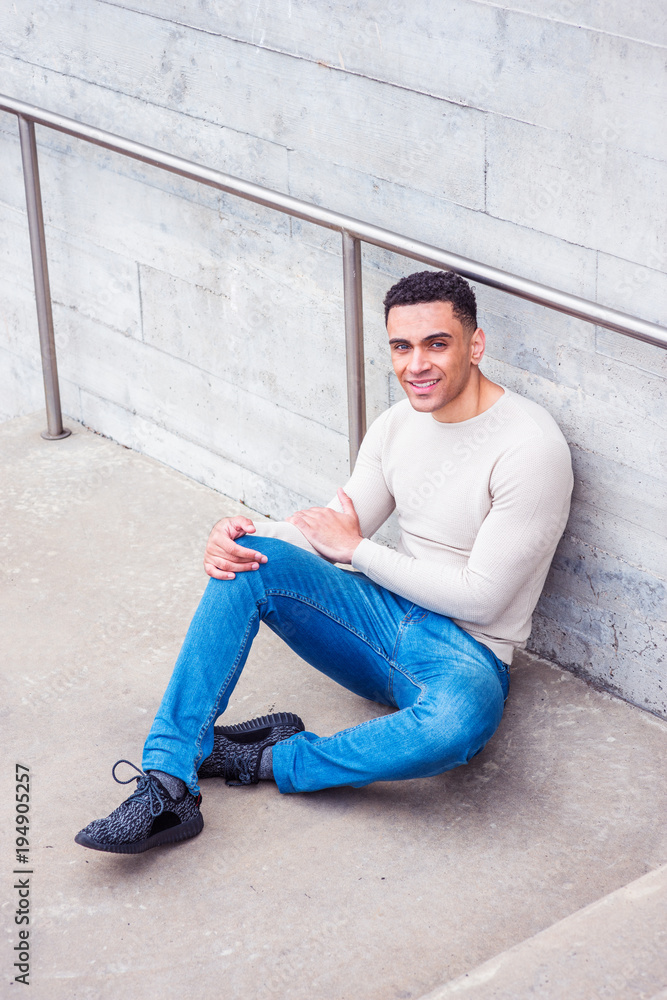 Young handsome American Man wearing light gray knit sweater, blue jeans, black sneakers, sitting on ground by wall in corner of street in New York, smiling, relaxing..