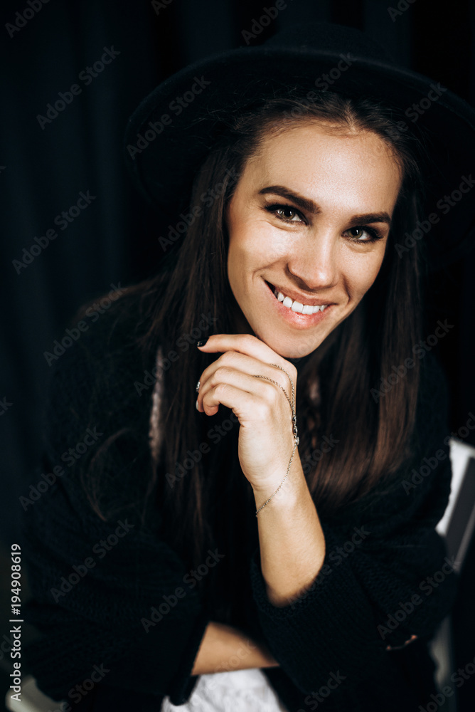 Emotional portrait of a beautiful smiling girl who sits on a black background in a black hat Emotional portrait of a beautiful smiling girl who sits on a black background in a black hat