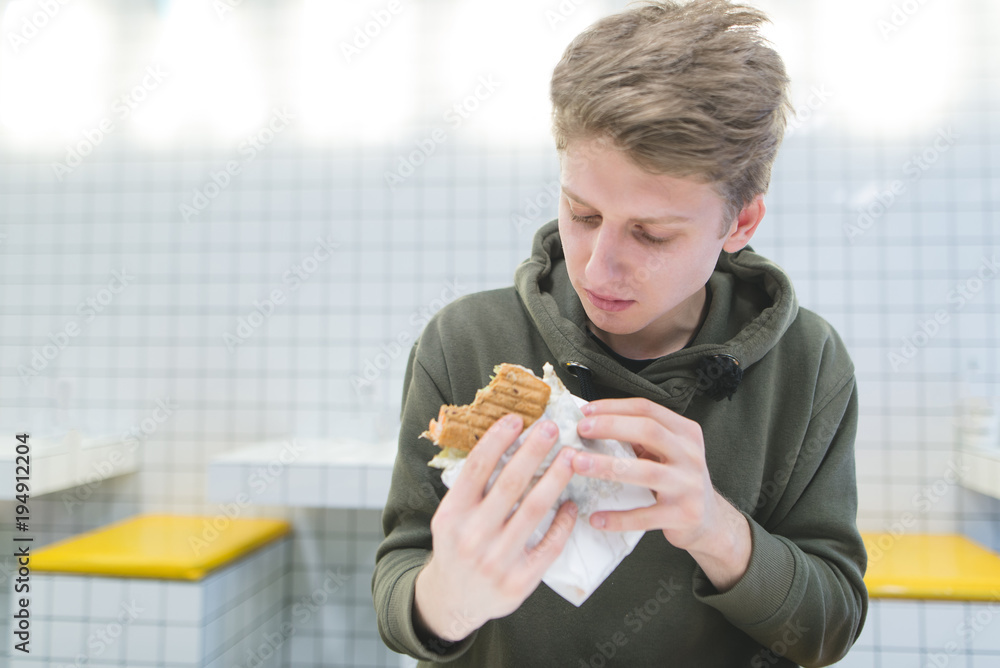 A student with a hungry look looks at a hamburger in his hands. Delicious fast food in a light restaurant with stylish design. A student with a hungry look looks at a hamburger in his hands. Delicious fast food in a light restaurant with stylish design.