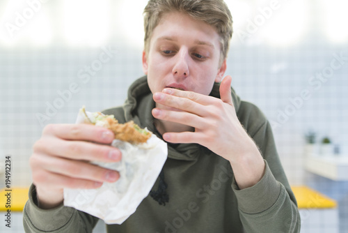 The student lickes his fingers and looks hungry looking at the saddle in his hands. Young man eats a hamburger in a fast food restaurant.