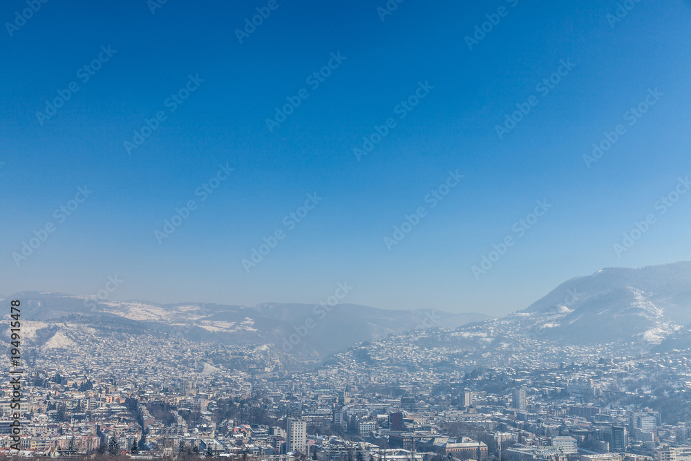 Aerial view of Sarajevo during a sunny winter afternoon, covered in snow. The historical center with its mosques and minarets can be seen in background