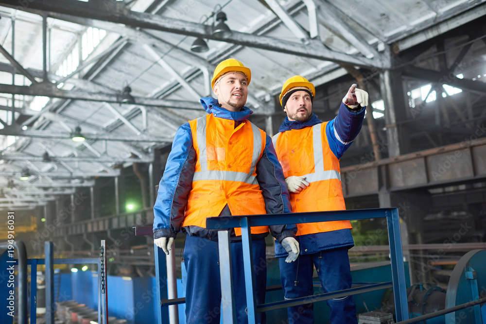 Team of hard-working technicians wearing uniform and hardhats looking at production department of modern plant while discussing faced issue, one of them pointing at something Team of hard-working technicians wearing uniform and hardhats looking at production department of modern plant while discussing faced issue, one of them pointing at something