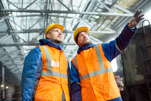 Low angle view of highly professional engineers wearing uniform and protective helmets standing at spacious production department of modern plant and discussing results of accomplished work.