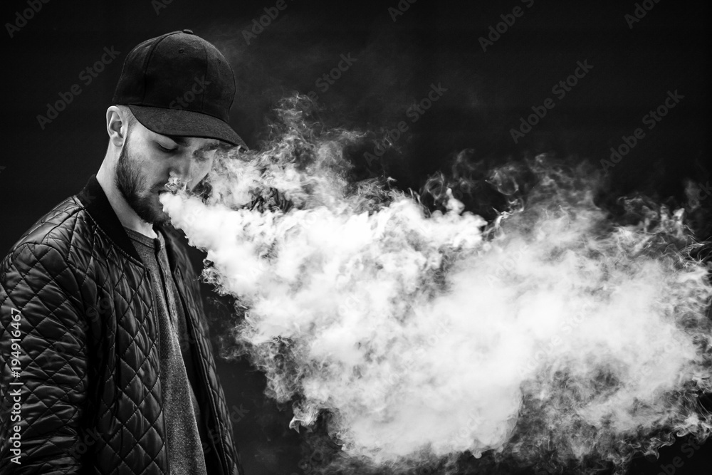 Vape man. Portrait of a handsome young white guy in a modern black cap  vaping and letting off puffs of steam from an electronic cigarette opposite  the Stock 写真 | Adobe Stock